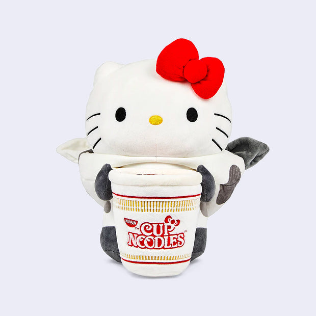 Plush version of Hello Kitty dressed in a cow suit with small horns and her red bow atop her head. She has her arms wrapped around a large Nissin Cup of Noodles. Her hood is off.