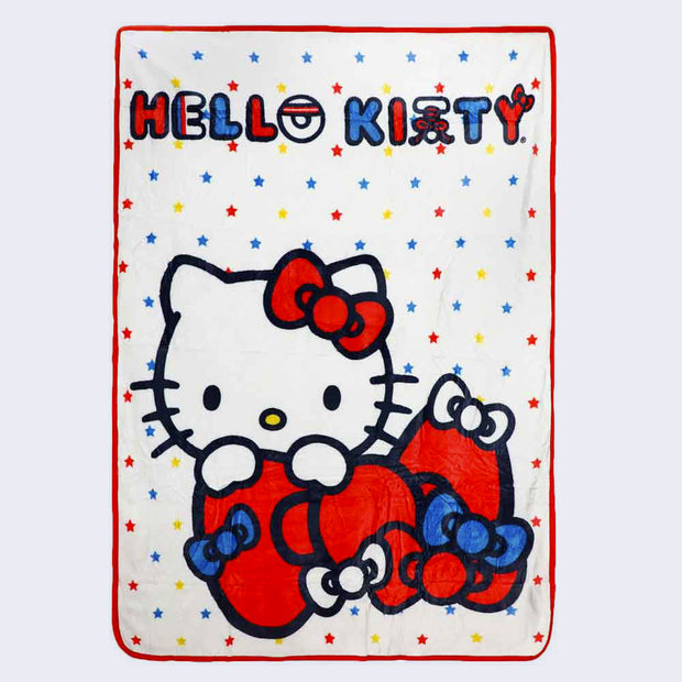 Spread out white plush blanket, with a red border and blue, yellow and red star pattern. "Hello Kitty" is written in stylized font at the top. An illustration of Hello Kitty, peeking over a large red bow with smaller bows attached to it is at the bottom center.
