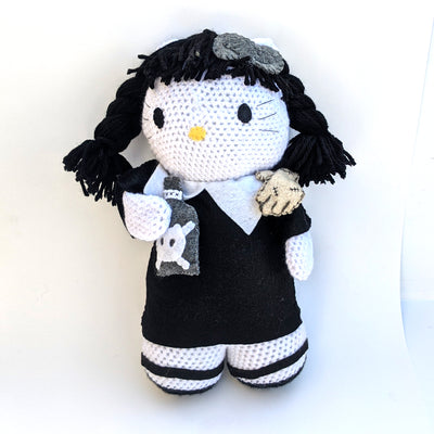 Crocheted plush doll of Hello Kitty, dressed as Wednesday Addams with dark braided hair, a black dress and a bottle of poison in her hands.