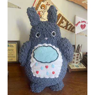 A tall dark gray Totoro inspired plush. On a white patch on its stomach is a raincloud with water droplets and hearts coming down from it.