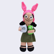 Crochet sculpture of Louise from Bob's Burgers, holding a mug of coffee that reads "I Will See You in Hell" and a green monster under her other arm.