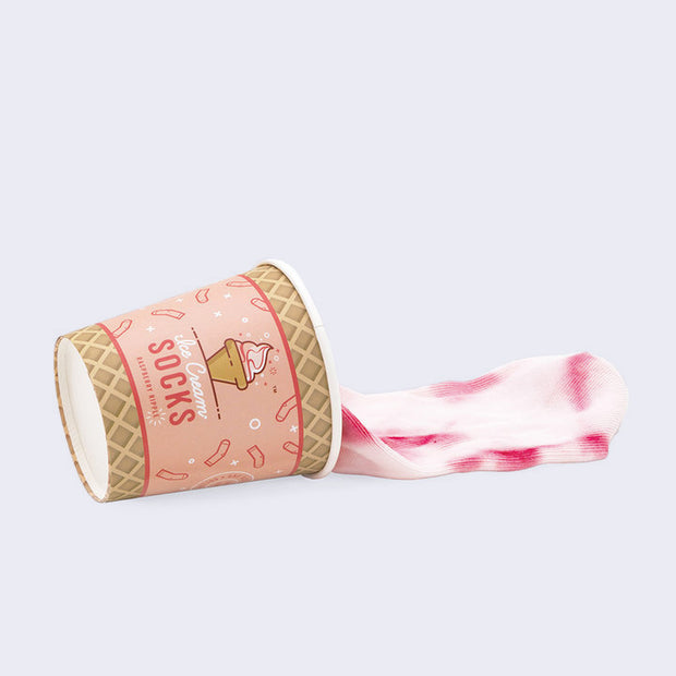 A tipped over cup with a pattern of an ice cream cone on it and a wrapping that reads "Ice Cream Socks." A pair of white and raspberry color tie dye socks spills out of the cup.