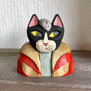 Whittled wooden sculpture of a black cat with pink ears and yellow eyes looking off to the side, dressed in a warm looking brown and red coat. Atop its head is a gray mouse. Sculpture sits flat as the cat is seen only from the chest up. 