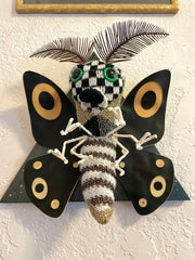 A sculpture of a moth, made out of mostly sweater like material, with checkered and stripe patterened body, green eyes, black wings with tan accents and thin leaf-like antennae. Mounted on a black wooden triangle.