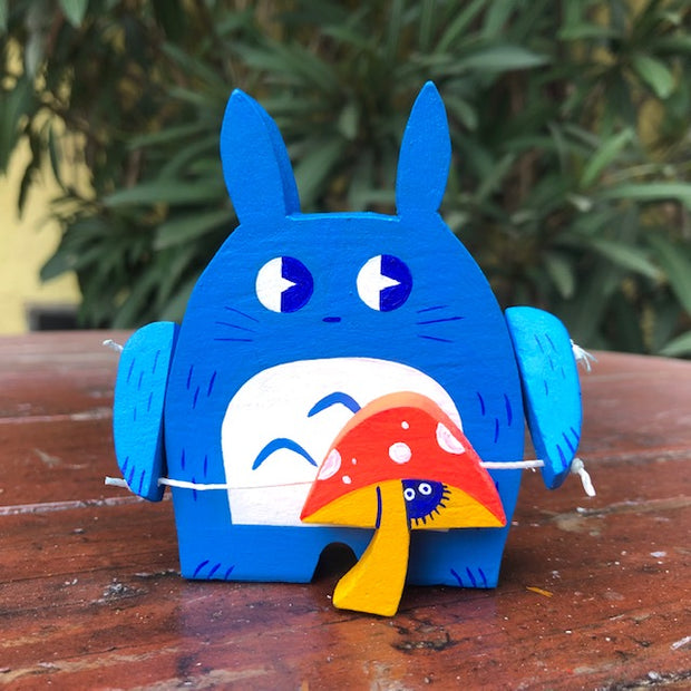 Wooden sculpture of a stylized small blue Totoro, looking off to the side and holding a red and yellow mushroom with a small dust sprite under the mushroom cap. Totoro's arms are attached with string, meaning they can move up and down freely.