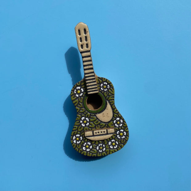 Stoneware 3D sculpture of a ukelele, with its body covered in a green leaf pattern with small white daisies. Body detailing and the neck are light tan, without any strings on the instrument.