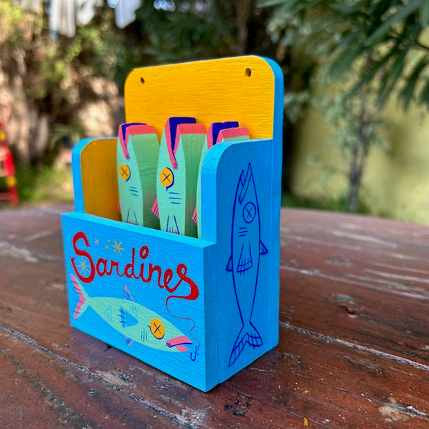 Painted wooden sculpture of a holder that says "sardines" on the front, with an illustration of a mint green sardine. Inside, are three mint green sardines with yellow x'd out eyes.