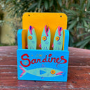 Painted wooden sculpture of a holder that says "sardines" on the front, with an illustration of a mint green sardine. Inside, are three mint green sardines with yellow x'd out eyes.