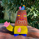 Die cut wooden sculpture of a tiki statue, with a human body. It has one hand extended out and the other as a thumbs up. Atop its head is a blue and pink bird.