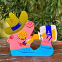 Die cut painted wooden sculpture of a pink man with a green cowboy hat and blue button up shirt. His eyes are yellow with red swirls and he drinks a bottle of yellow liquid with a blue moon label on it.