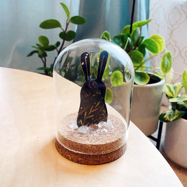Glass dome with a cork base with a cut out shape of a simple black rabbit with gold painted pattern details and accents. Around the bunny appears to be melted snow, made out of resin.