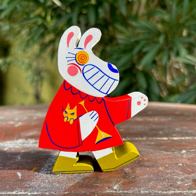 Die cut wooden sculpture of a white rabbit with yellow swirl pattern eyes and a large tooth grin, wearing a red collared dress with olive green boots, holding a brass horn in its hand and a small yellow kitten in the crook of its arm.