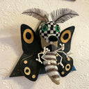 A sculpture of a moth, made out of mostly sweater like material, with checkered and stripe patterened body, green eyes, black wings with tan accents and thin leaf-like antennae.  Mounted on a black wooden triangle.