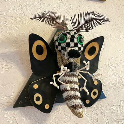 A sculpture of a moth, made out of mostly sweater like material, with checkered and stripe patterened body, green eyes, black wings with tan accents and thin leaf-like antennae.  Mounted on a black wooden triangle.