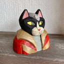 Whittled wooden sculpture of a black cat with pink ears and yellow eyes looking off to the side, dressed in a warm looking brown and red coat. Atop its head is a gray mouse. Sculpture sits flat as the cat is seen only from the chest up.