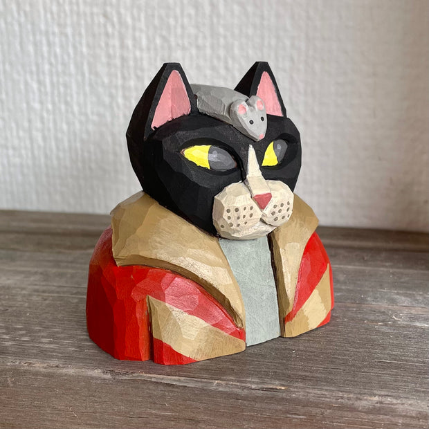 Whittled wooden sculpture of a black cat with pink ears and yellow eyes looking off to the side, dressed in a warm looking brown and red coat. Atop its head is a gray mouse. Sculpture sits flat as the cat is seen only from the chest up.