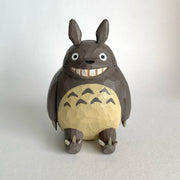 Whittled wooden sculpture of Totoro, sitting and smiling. His body is round and his ears are pointed, with sharp claws on his hands and feet.