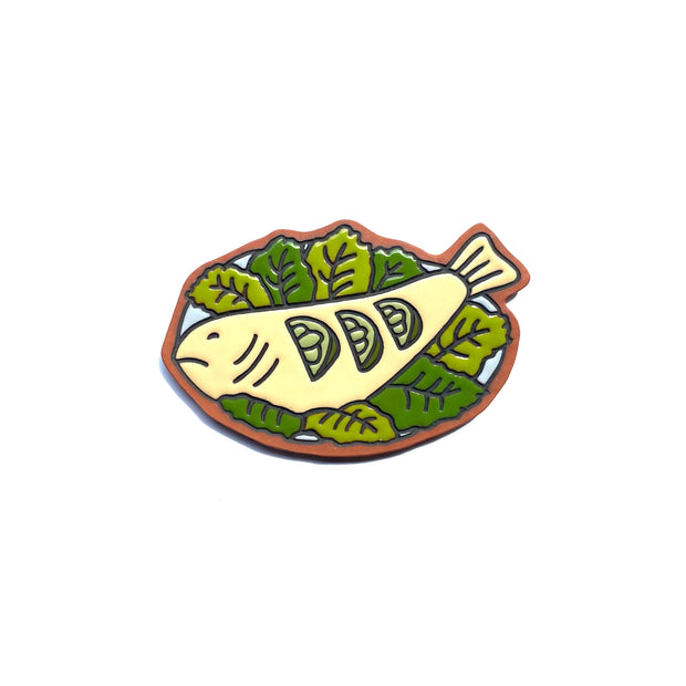 Earthenware die cut tile of a yellow fish served on a plate with many green leaves as garnish and 3 lime slices atop its body. 