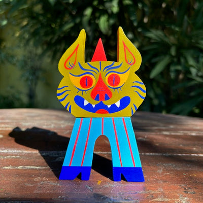 Die cut painted wooden sculpture of a small green pig-like gremlin, with a smiling face and tiger stripes on its face. It wears a pointy red hat, blue pin striped pants and blue boots.