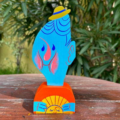Die cut painted wooden sculpture of the bust of a blue man, with squiggly features and worried eyes. He holds a small smiling half sun in his hands against his chest.
