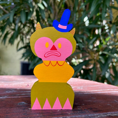 Die cut painted wooden sculpture of a pink frowning clown creature, with small green horns and a blue top hat.