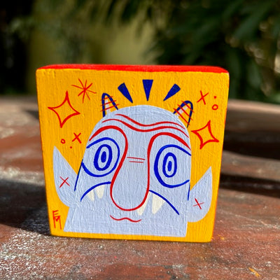 Painted wooden block, bright yellow with an illustration of a curious looking lavender devil on it with a long nose. 