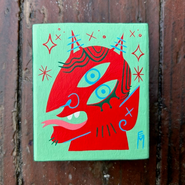 Small wooden block, painted mint green with an illustration of a red devil with two eyes. Its forked tongue is out of its mouth, with sparkles around its head.