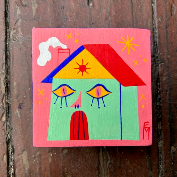 Coral pink painted wooden square, with an illustration of a mint green house with a primary colored roof. The house has mascara'd eyes and a nose, with the red door as the mouth.