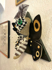 A sculpture of a moth, made out of mostly sweater like material, with checkered and stripe patterened body, green eyes, black wings with tan accents and thin leaf-like antennae. Mounted on a black wooden triangle.