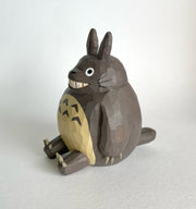 Whittled wooden sculpture of Totoro, sitting and smiling. His body is round and his ears are pointed, with sharp claws on his hands and feet. Side angle.