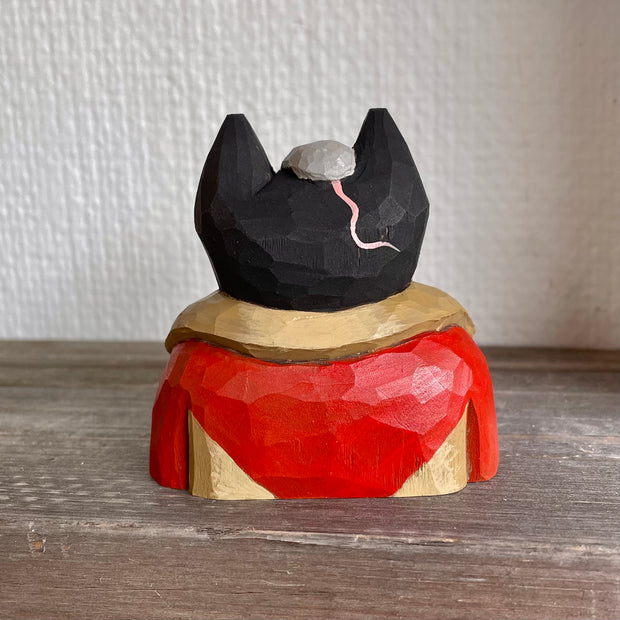 Back view of whittled wooden sculpture of a black cat, visible only from the chest up. Its dressed in a warm looking red and brown coat with a gray mouse atop its head, the pink tail showing from behind.