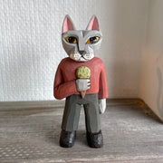 Whittled wooden sculpture of a surly looking gray cat with yellow eyes, standing on two legs like a human. They are dressed in a red long sleeve and gray slacks with black boots and hold an ice cream cone with a scoop of green ice cream.