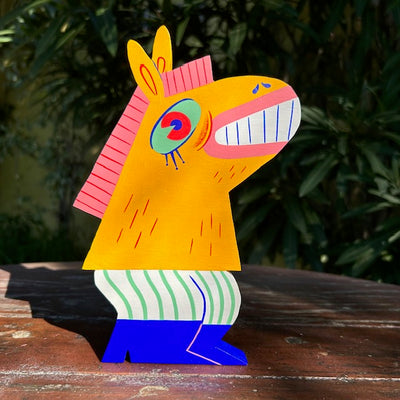 Die cut painted wooden sculpture of a smiling yellow horse, with a large green eye and pink mane. Its standing on 2 legs, like a human and wearing pin striped pants and blue boots.
