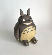 Whittled wooden sculpture of Totoro, sitting and smiling. His body is round and his ears are pointed, with sharp claws on his hands and feet.
