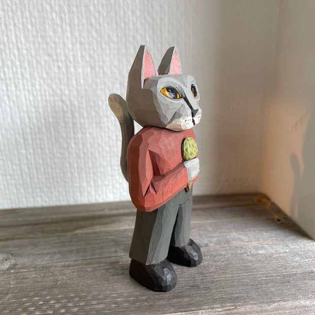 Whittled wooden sculpture of a surly looking gray cat with yellow eyes, standing on two legs like a human. They are dressed in a red long sleeve and gray slacks with black boots and hold an ice cream cone with a scoop of green ice cream. It has an upright, long tail.