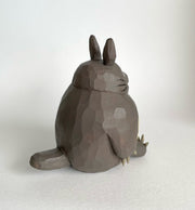 Whittled wooden sculpture of Totoro, sitting and smiling. His body is round and his ears are pointed, with sharp claws on his hands and feet. Back view.