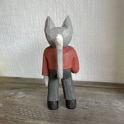 Back view of a whittled wooden gray cat sculpture, standing up on 2 legs like a human. They are dressed in a red long sleeve and gray slacks with black boots and hold an ice cream cone with a scoop of green ice cream. It has an upright, long tail.