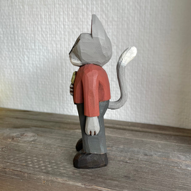 Whittled wooden sculpture of a surly looking gray cat with yellow eyes, standing on two legs like a human. They are dressed in a red long sleeve and gray slacks with black boots and hold an ice cream cone with a scoop of green ice cream. It has an upright, long tail.