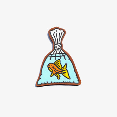 Earthenware die cut tile of a tied plastic bag holding a goldfish in light blue water. The goldfish has yellow fins and looks up, with a slight smiling expression. 