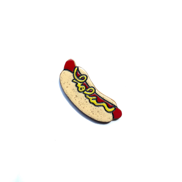 Stoneware die cut tile of a red hot dog in a speckled light tan bun with cursive yellow mustard that reads "hola."