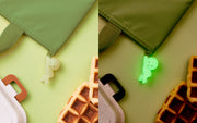 Smiski attached to a zipper with their arms extended and knees slightly lifted, in a "hanging on" position. One image is of Smiski regularly and the other is of them glowing in the dark.