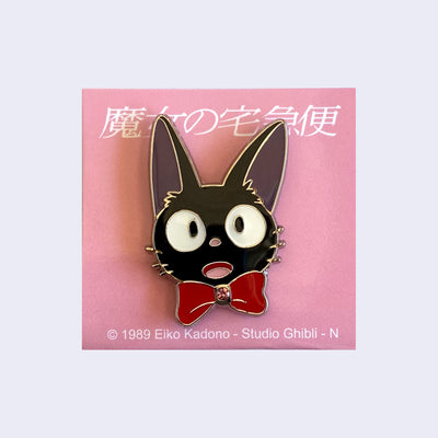 Enamel pin of a close up Jiji head, with a surprised and happy expression, wearing a big red bow that has a pink gem in the middle of it. 