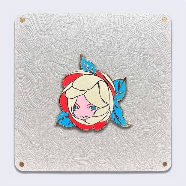 Silver outlined enamel pin of a blooming chrysanthemum like flower, with many grayish cream colored petals and bright red inner petals and greenish blue leaves. A small pink face peeks out from inside the bulb. Pin is on an ornate patterned white backing card.