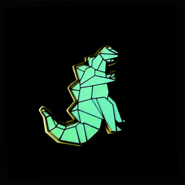 Enamel pin of an origami kaiju monster, standing and facing the right with spikes on its back and a large tail. Gold outlining. Pin is glowing in the dark.