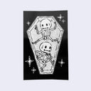 Black vertical rectangle sticker with a simplistic black and white illustration of two skeletons in a coffin, one resting its head on the back of its hand and the other with a peace hand sign. 