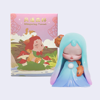 Vinyl figure of a cherub like girl with long semi transparent teal blue to purple ombre hair and a long puffy blue dress, she stands with her eyes closed and a solemn expression. One goldfish hangs from her head and the other appears over her shoulder. She's positioned in front of Whisper Forest blind box packaging.