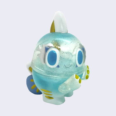Small soft vinyl figure of a cartoon fish creature, with large dark blue eyes and a small pink smile. It has large rounded spikes on its head and body and it is teal blue on top with a bluish clear glitter middle and blue feet with gold accents.