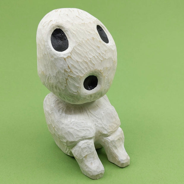 Whittled wooden sculpture of a simplistically shaped white creature, sitting with its hands on its knees. It has a large head to its small body, with large black oval shaped eyes and an open black mouth.