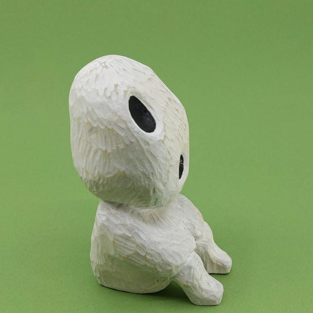 Whittled wooden sculpture of a simplistically shaped white creature, sitting with its hands on its knees. It has a large head to its small body, with large black oval shaped eyes and an open black mouth. Side view.