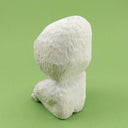 Back view. Whittled wooden sculpture of a simplistically shaped white creature, sitting with its hands on its knees. It has a large head to its small body, with large black oval shaped eyes and an open black mouth.